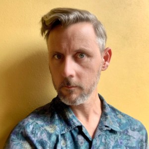 Photo of a white man in a blue shirt against a yellow background