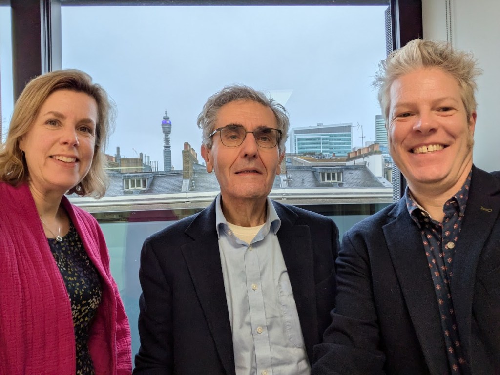 Three people, 2 men and a woman stood in front of the London skyline smiling at the camera